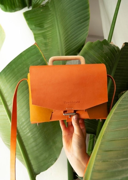 Imagen de bolso de color naranja, soportada por una mano en un fondo de hojas de plantas verdes